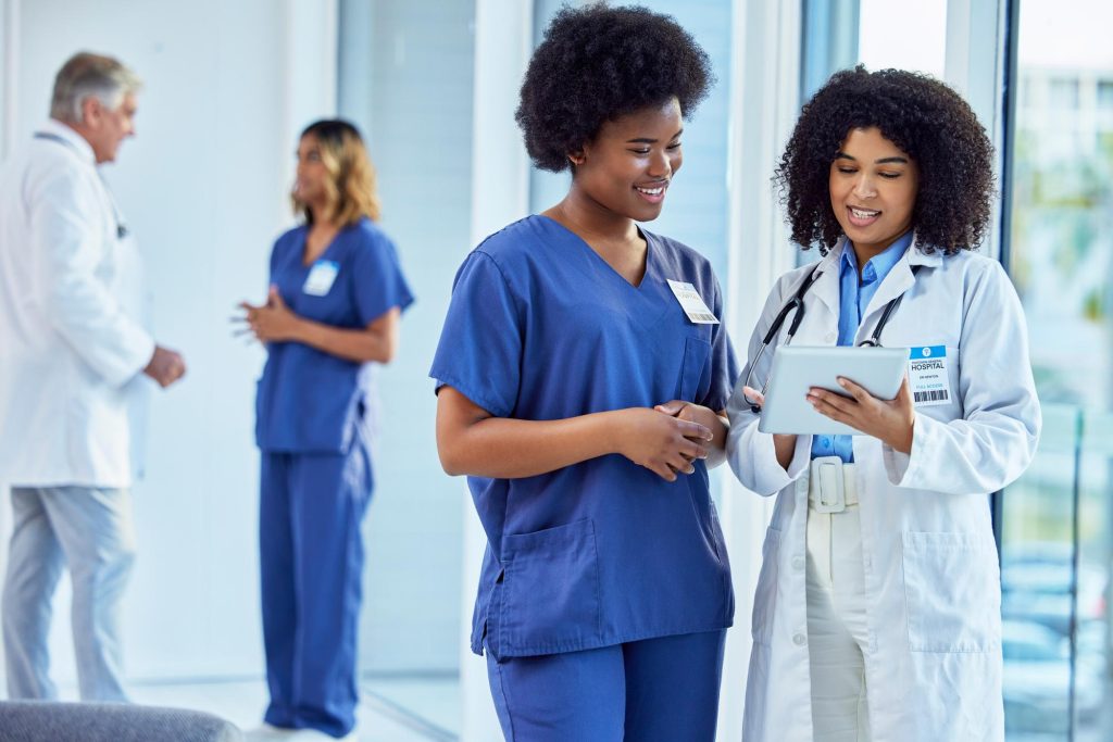 Two clinicians, one in scrubs and one in a white coat, looking at a tablet together while colleagues talk behind them.