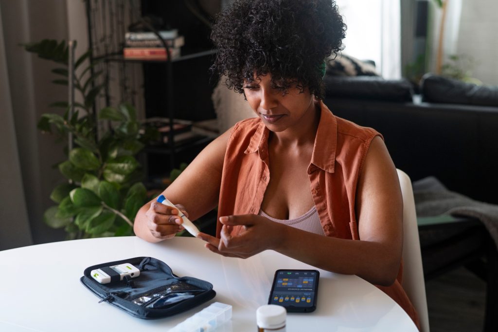 Woman at a table pricking her fingertip with a lancing pen, with an open blood glucose test kit, a pill bottle and a phone app in front of her.