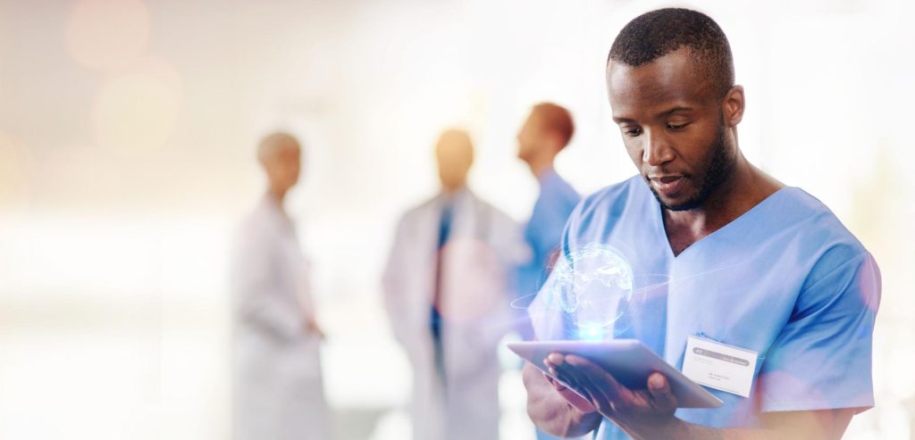 Clinician in light blue scrubs reading a tablet that projects a glowing globe graphic, with colleagues out of focus behind him.
