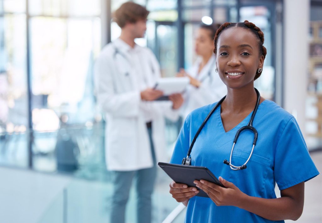 Nurse in blue scrubs with a stethoscope, holding a tablet, two colleagues in conversation behind her.