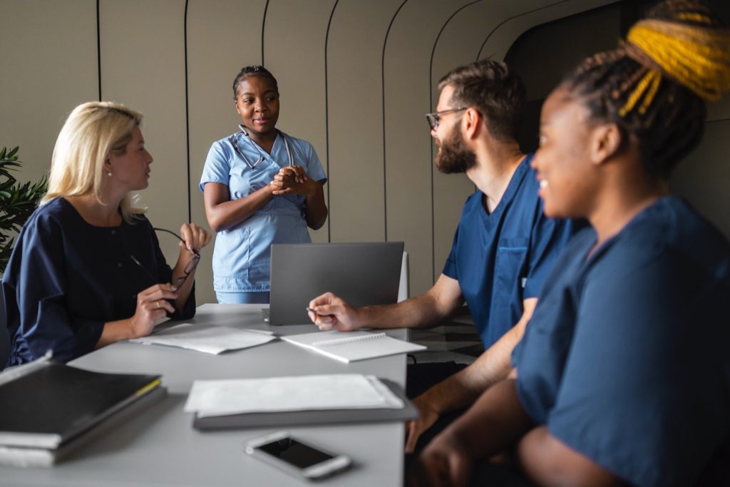Four colleagues around a meeting table, one standing in scrubs speaking to the group.